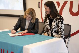 Dr. Lorrie Clemo, President of D'Youville University Signs the Okanagan Charter at a table. Paige Schultze, LCMHC, D'Youville University Clinical Mental Health Counselor, sits next to her at the table.
